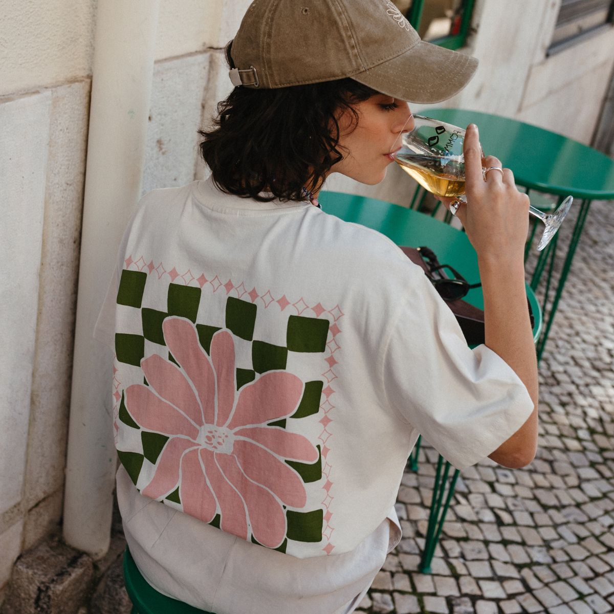 Back view of a woman wearing an off-white TBCo Cotton t-shirt with bold pink floral and green checkerboard graphic, sipping white wine at an outdoor café with green tables and a cobbled street.