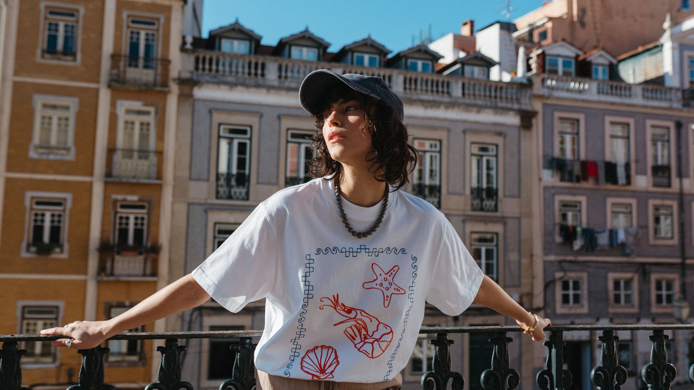 TBCo Cotton T-Shirt in Shrimp, a relaxed-fit white tee featuring a bold red shrimp, starfish, and seashell print, styled casually with a cap and necklace against a European city backdrop.