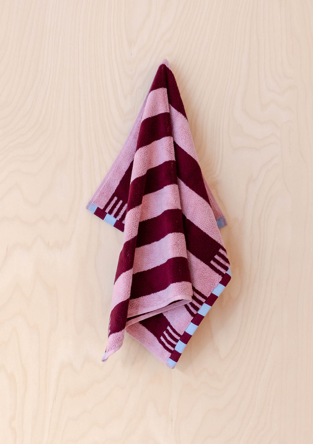 TBCo Terry Cotton Towels in Burgundy Stripe hanging against pale wood wall showing pink and burgundy stripes with blue checkerboard edges