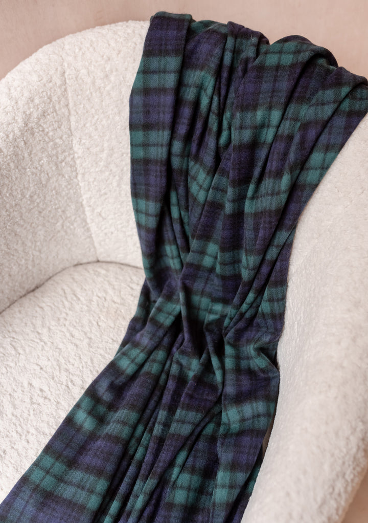 Soft TBCo Cashmere Blanket in Black Watch Tartan draped over a cream textured chair showcasing elegant warmth and style