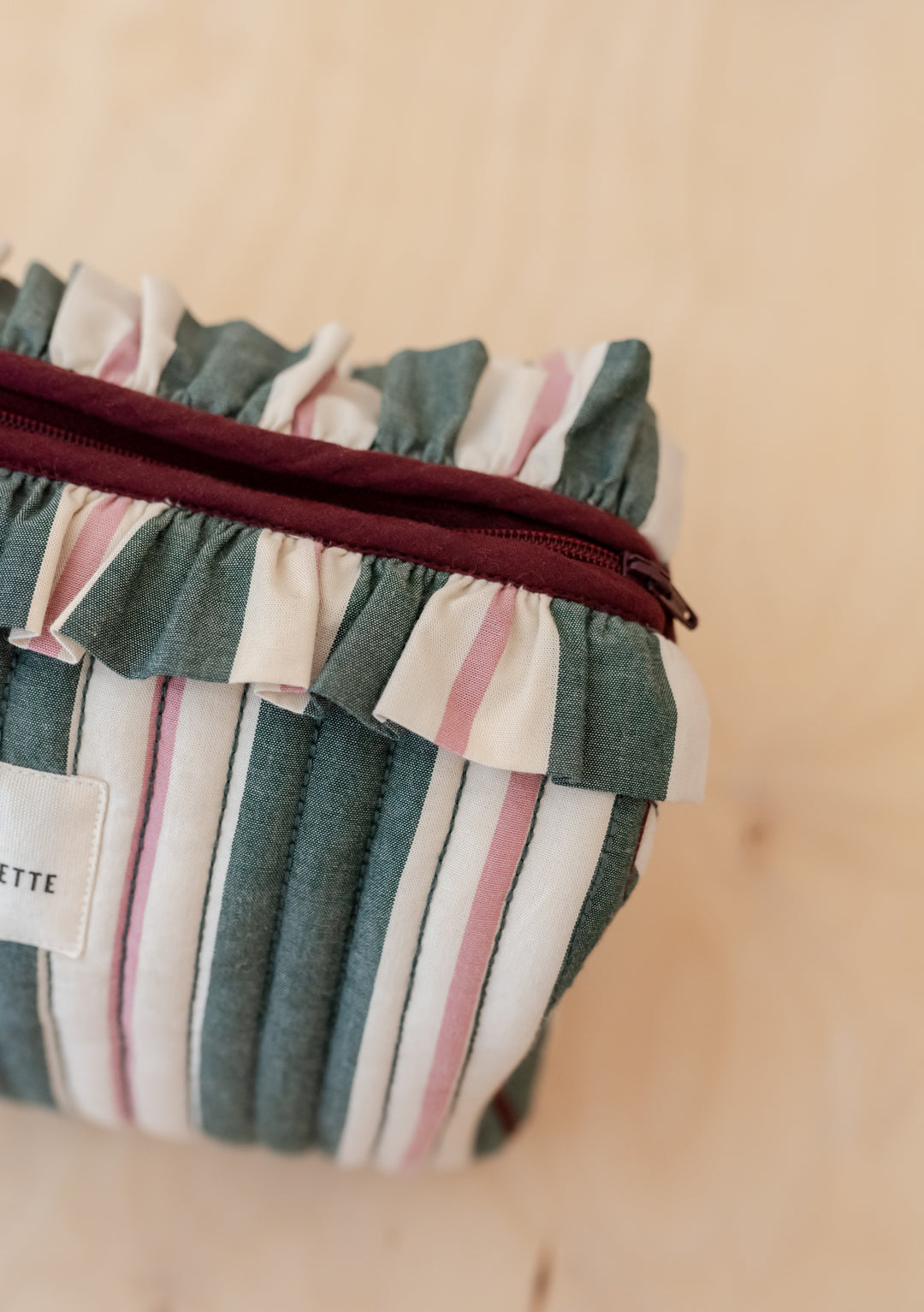 TBCo x Glassette Cotton Wash Bag in Green Stripe, featuring quilted fabric, burgundy zipper, ruffled trim, and multiple interior compartments on a light wood surface.